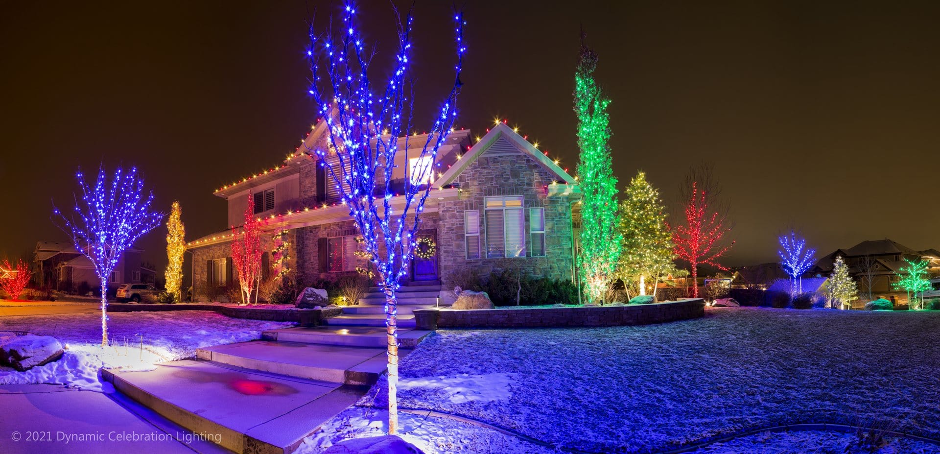 House and yard decorated with colorful holiday lights on trees and roof, snow covering the ground.