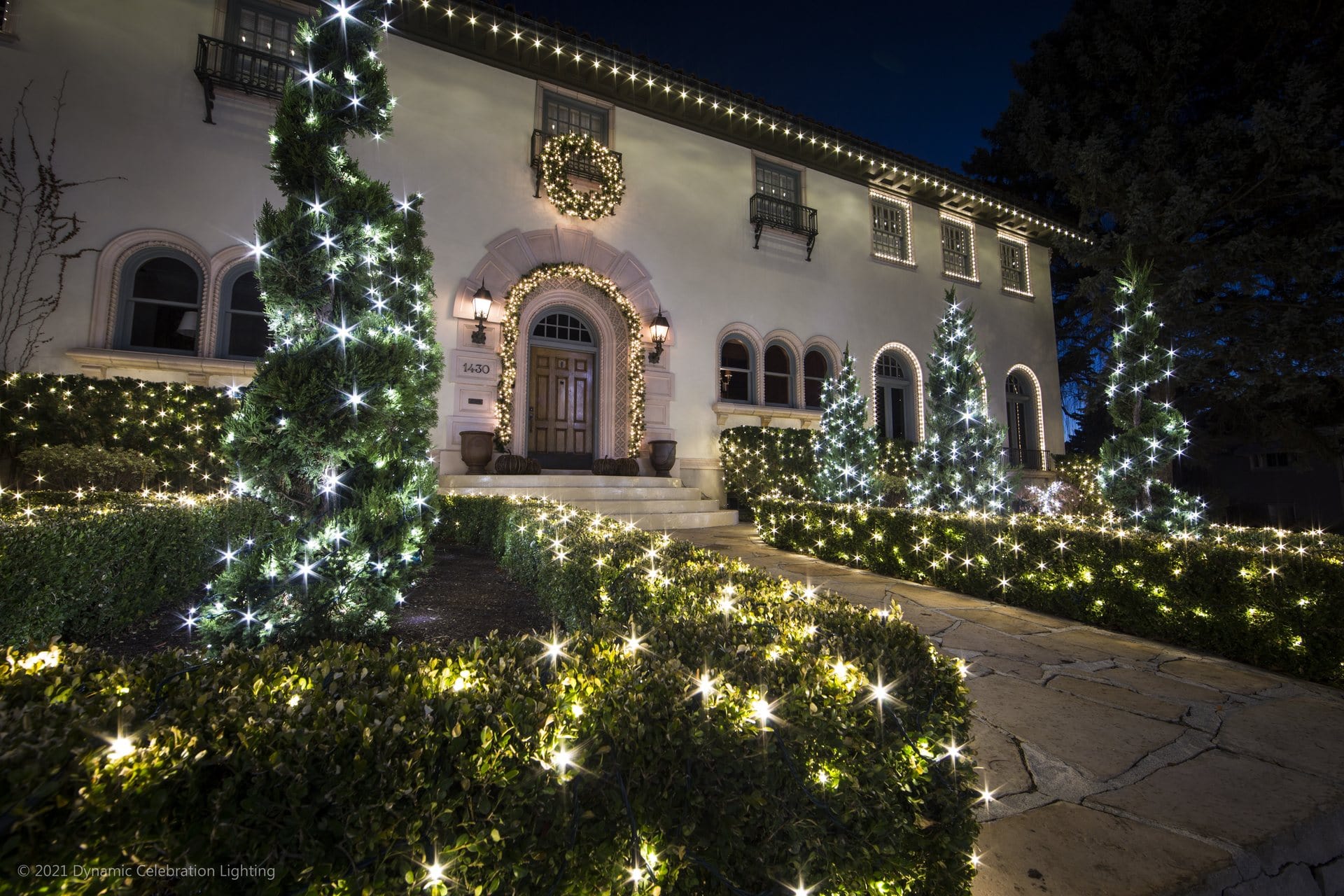 A house decorated with white holiday lights outlining bushes, trees, windows, and a wreath above the door.