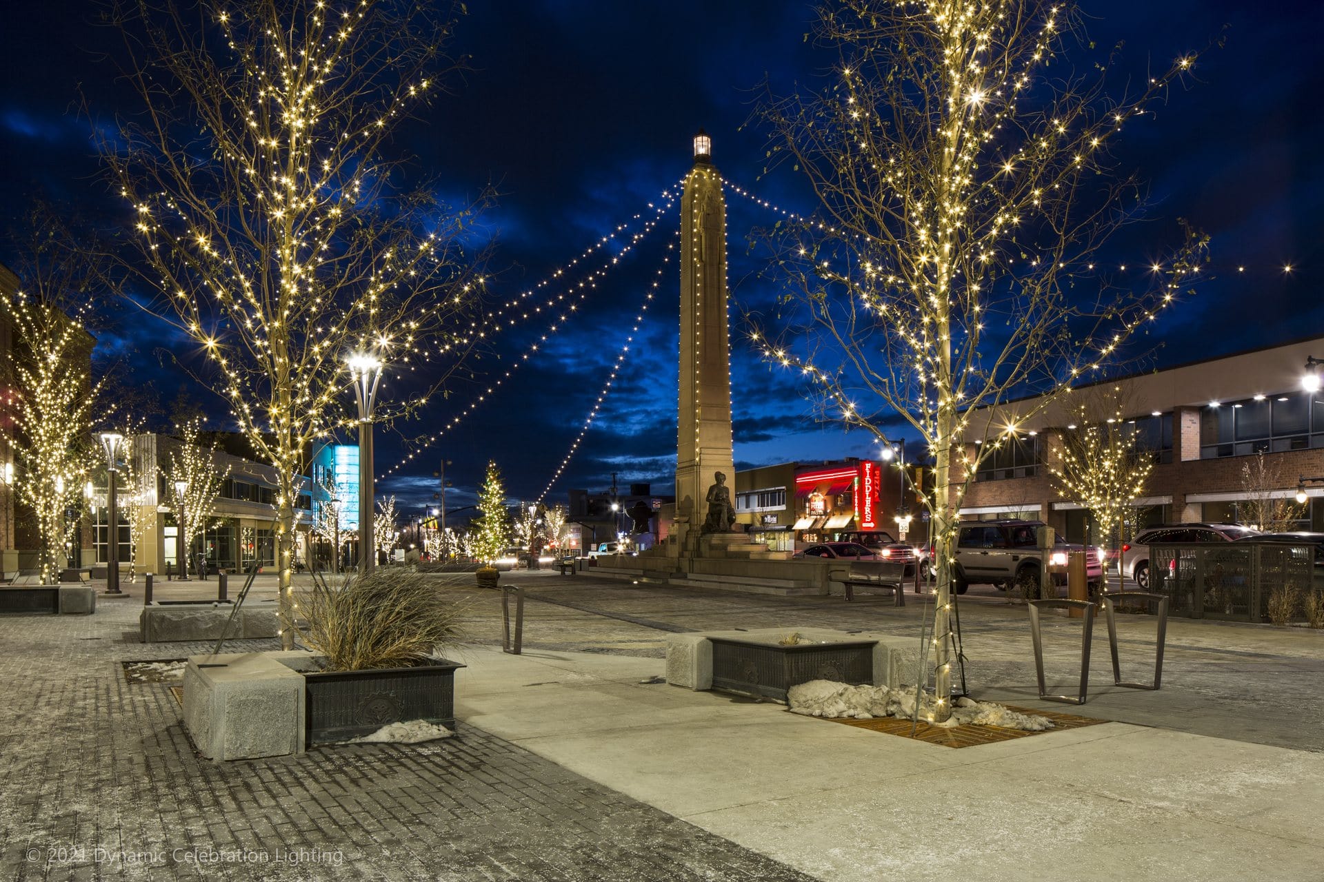 City street at night with trees and lampposts wrapped in string lights; a tall monument in the background.