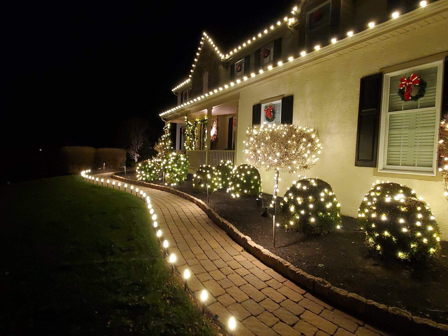 House and bushes decorated with white holiday lights; wreaths on doors and windows; brick walkway.