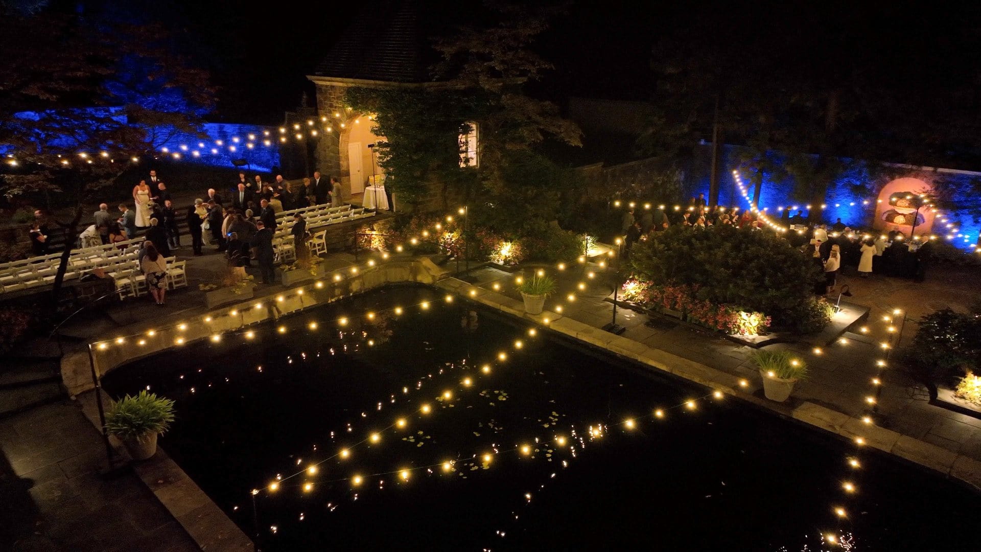 Outdoor night wedding with string lights over a pond, guests gathered near a stone building and garden.