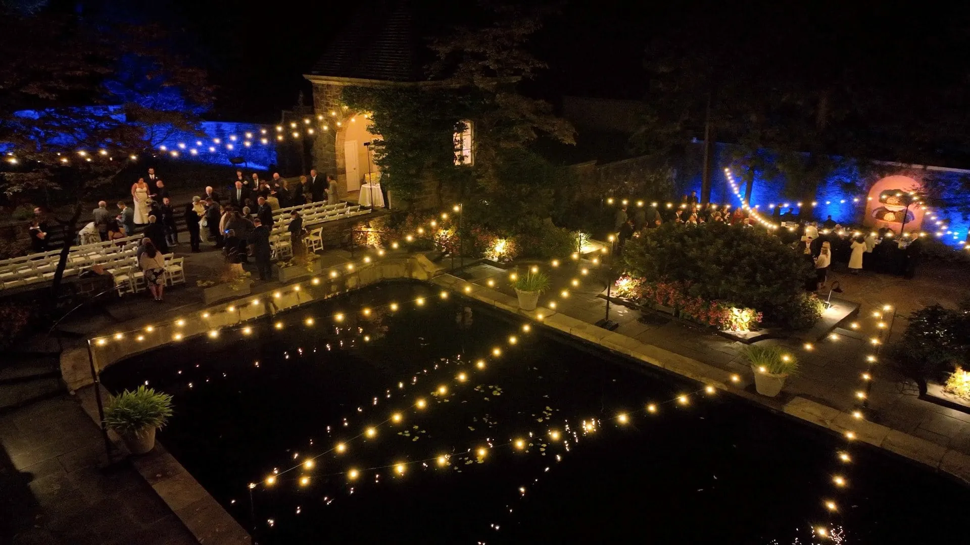 Nighttime outdoor wedding with string lights over a pond and guests gathered near trees and a stone building.