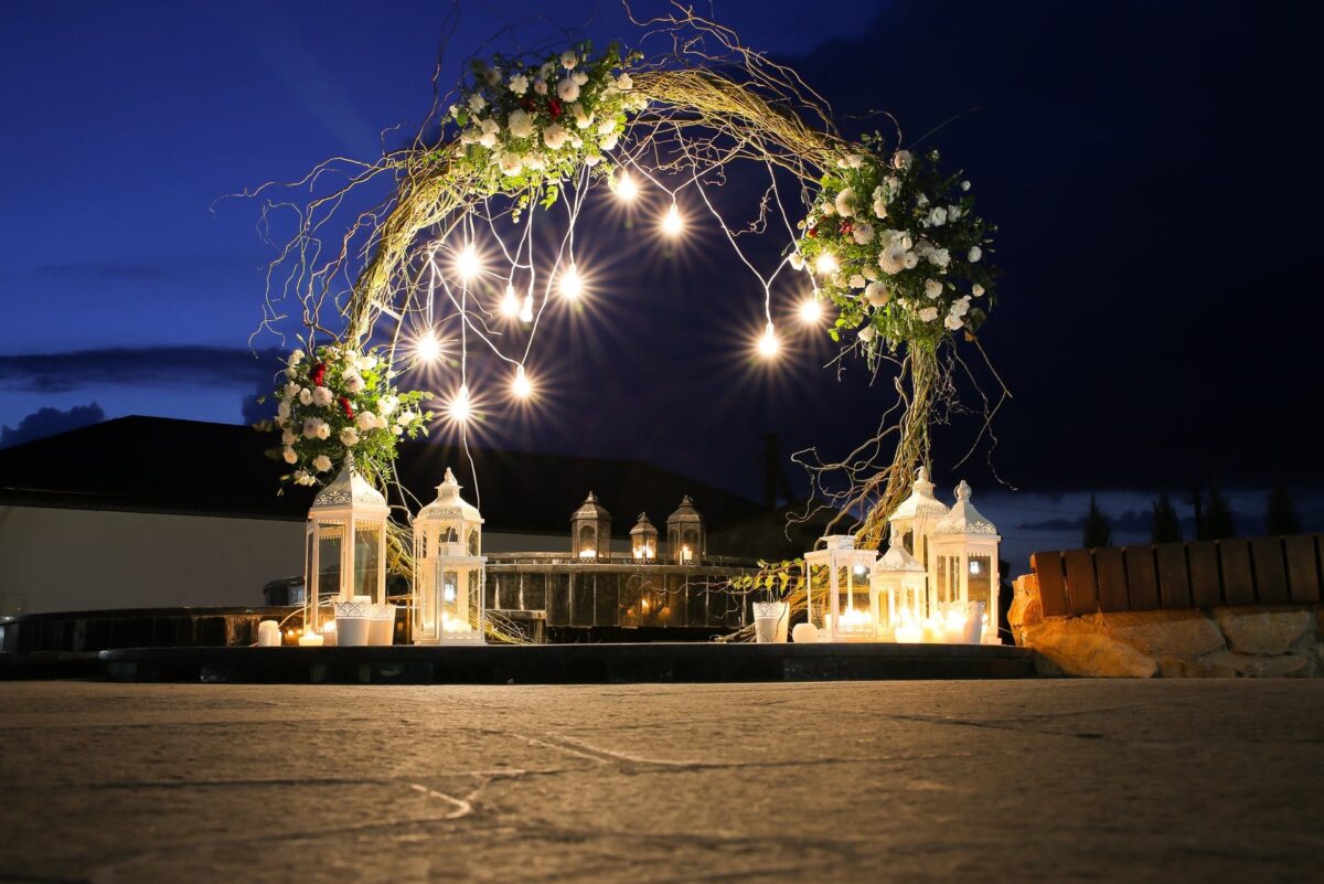 A floral arch with hanging lights and lanterns is set up outdoors at dusk, creating a romantic scene.