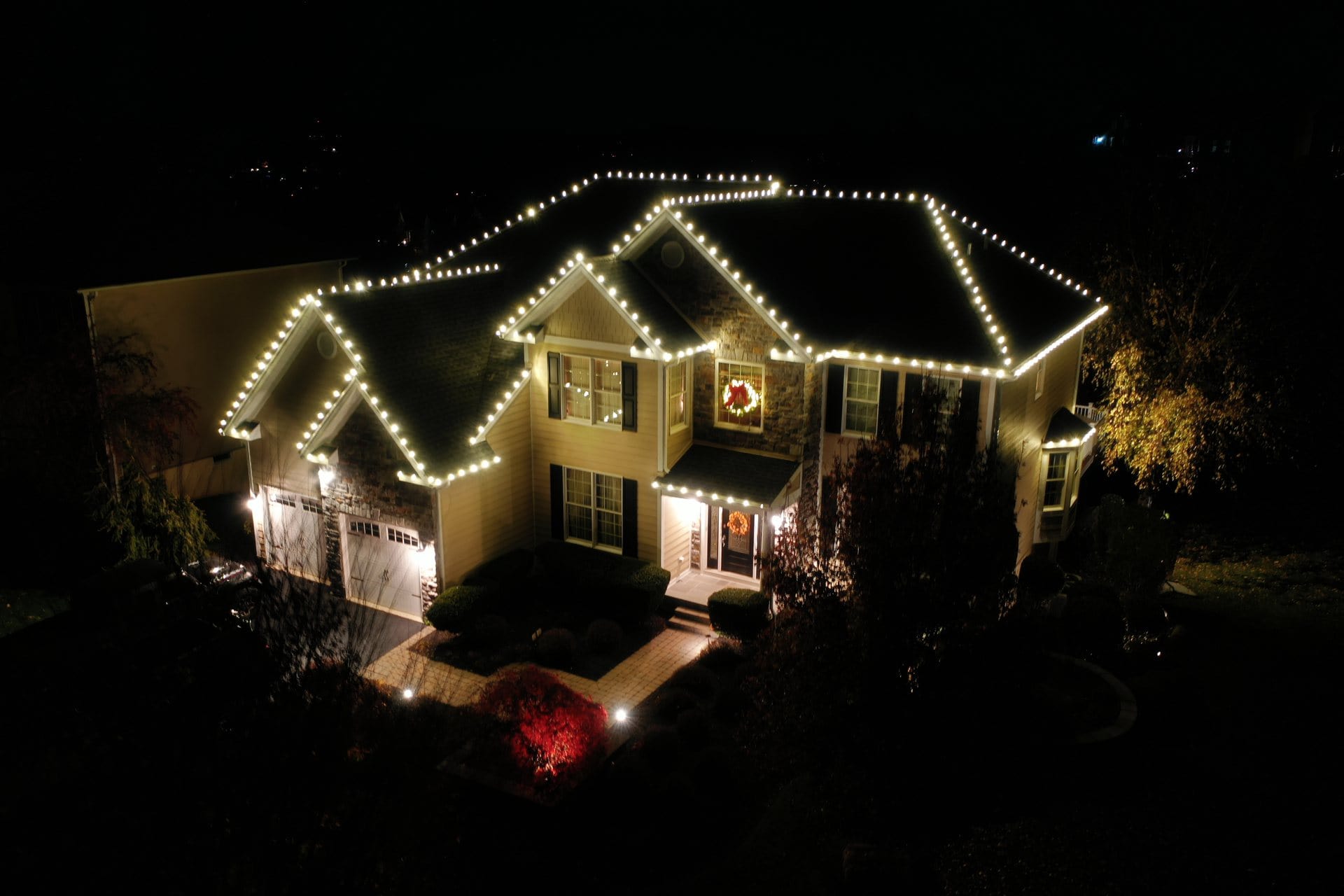 A two-story house at night, decorated with white holiday lights and a wreath on the front door.