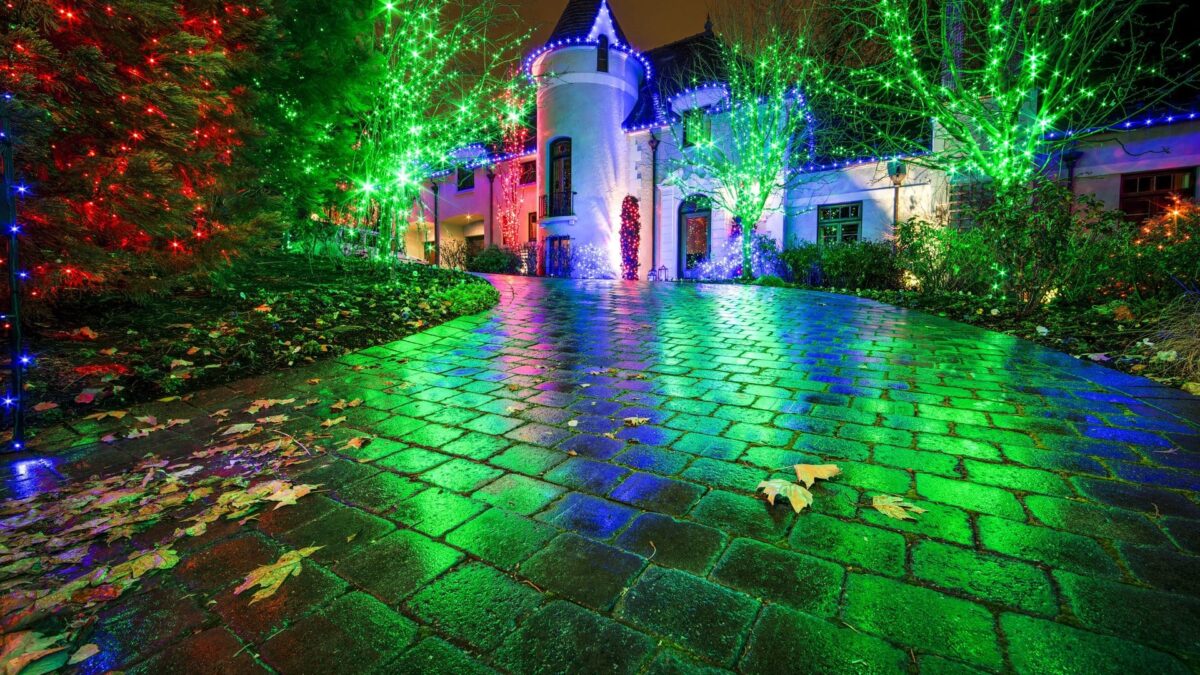 A house and driveway lit up with colorful holiday lights at night, reflecting on wet cobblestones.