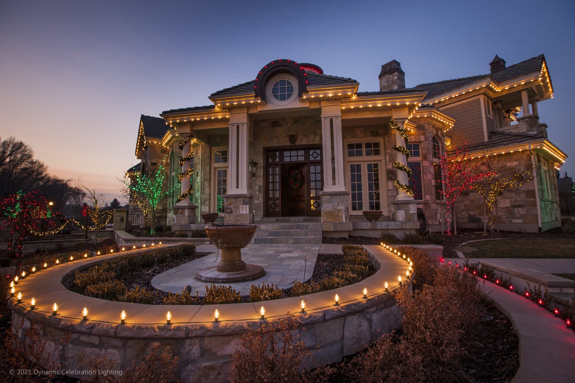 A stone house decorated with Christmas lights at twilight, featuring a lit fountain in the foreground.
