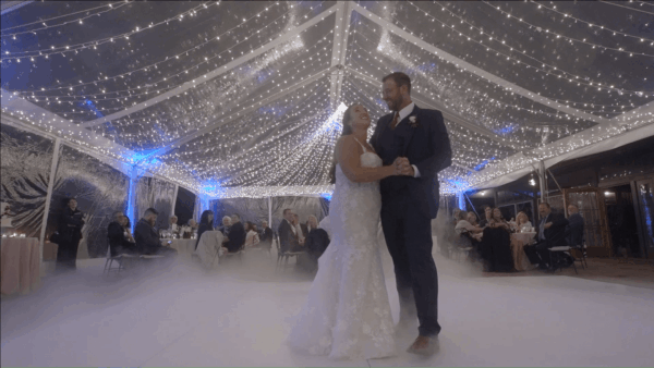 Bride and groom share a first dance under string lights at a wedding reception with guests seated nearby.