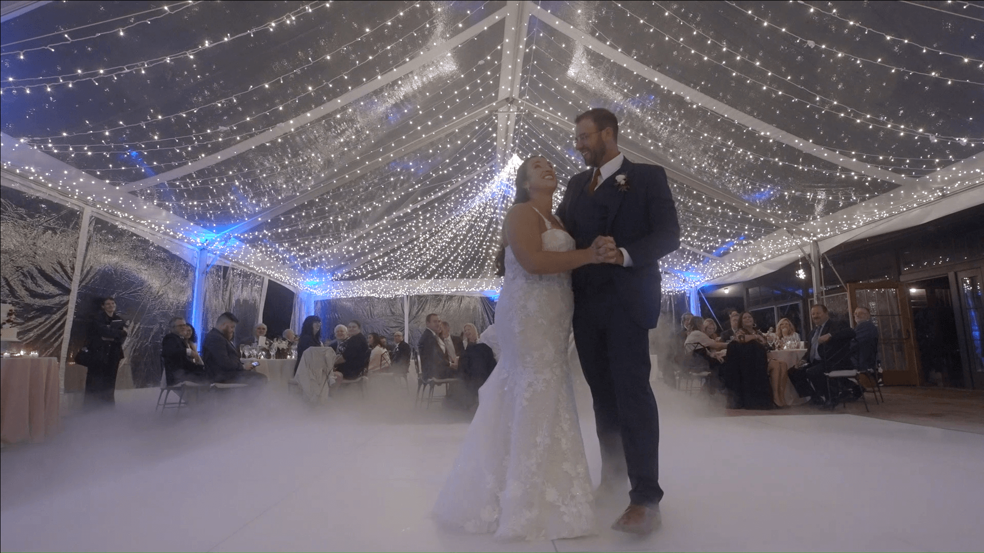 Bride and groom share a first dance under string lights at a wedding reception with guests seated nearby.