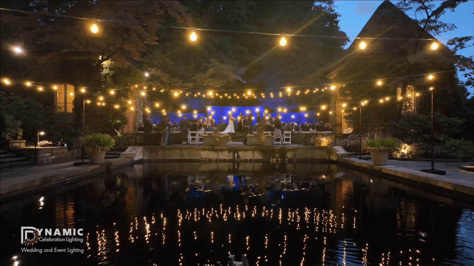 A wedding reception at night with string lights and reflections over a pond, guests seated in the background.