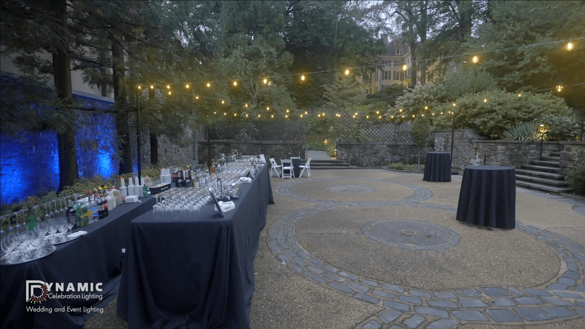 Outdoor event setup with string lights, cocktail tables, and a bar under trees in the evening.