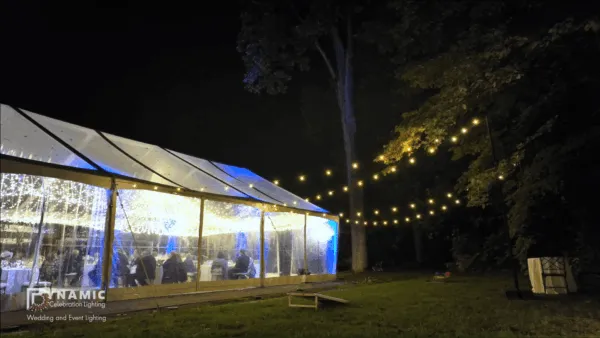 Outdoor wedding tent at night, lit with string lights, surrounded by trees and grass.