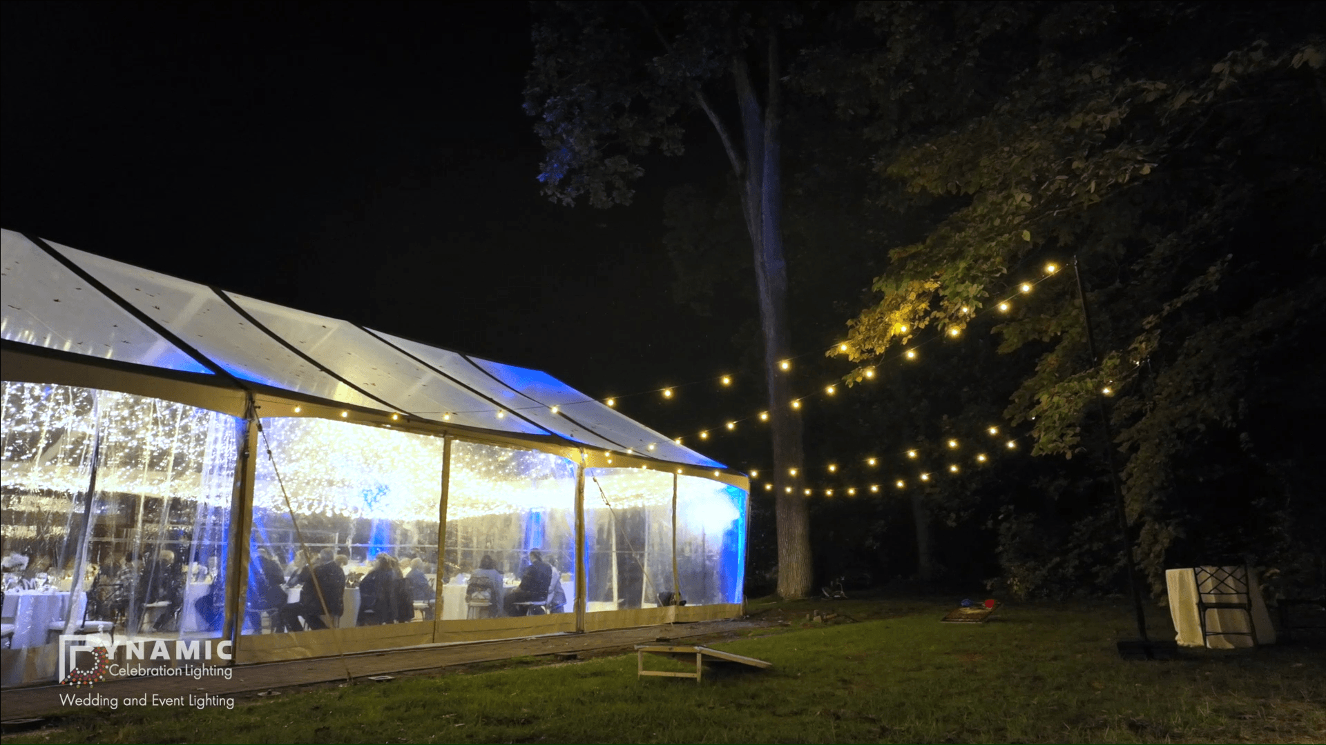 Outdoor wedding tent at night, lit with string lights, surrounded by trees and grass.
